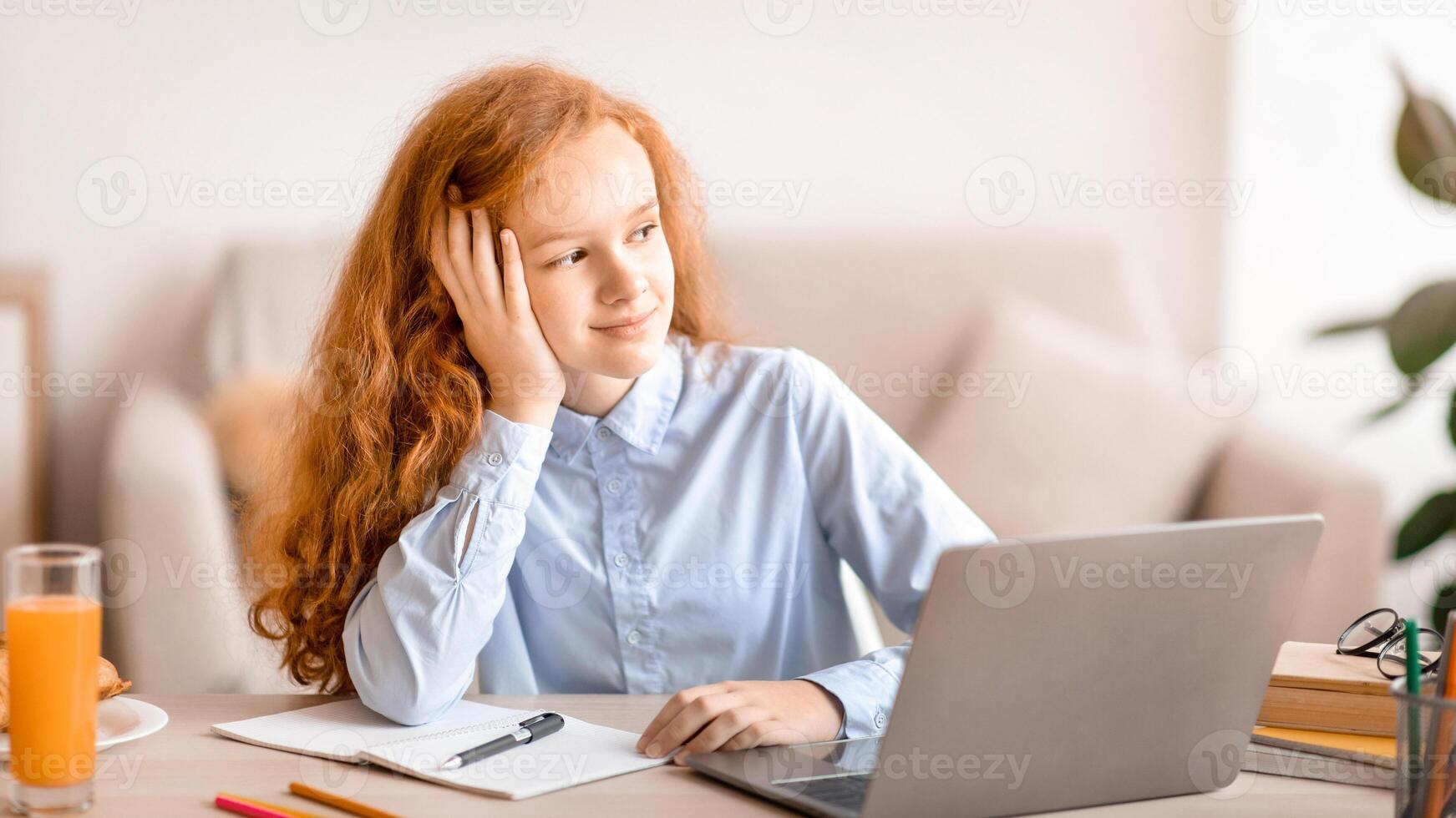 Portrait of smiling red-haired girl sitting at desk with laptop computer and looking away at window, thinking and dreaming, doing homework, studying online at home in her bedroom. Remote Learning photo
