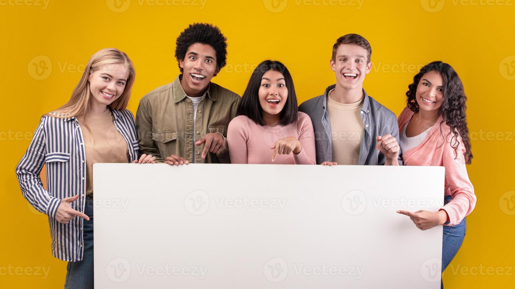 Group of friends laughing, sharing good and positive mood. Happy young multiracial students with open mouths hold banner with blank space, isolated on orange background, studio shot, panorama photo