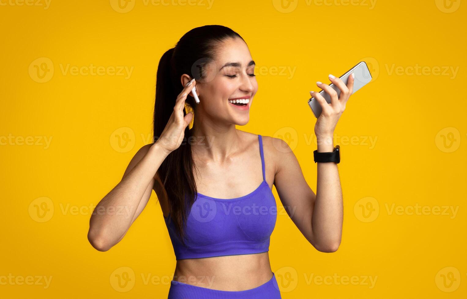Music For Workout. Portrait Of Smiling Young Slim Lady Wearing Wireless Earphones, Enjoying Favorite Song And Playlist For Training, Holding Smartphone Isolated Over Orange Studio Background photo