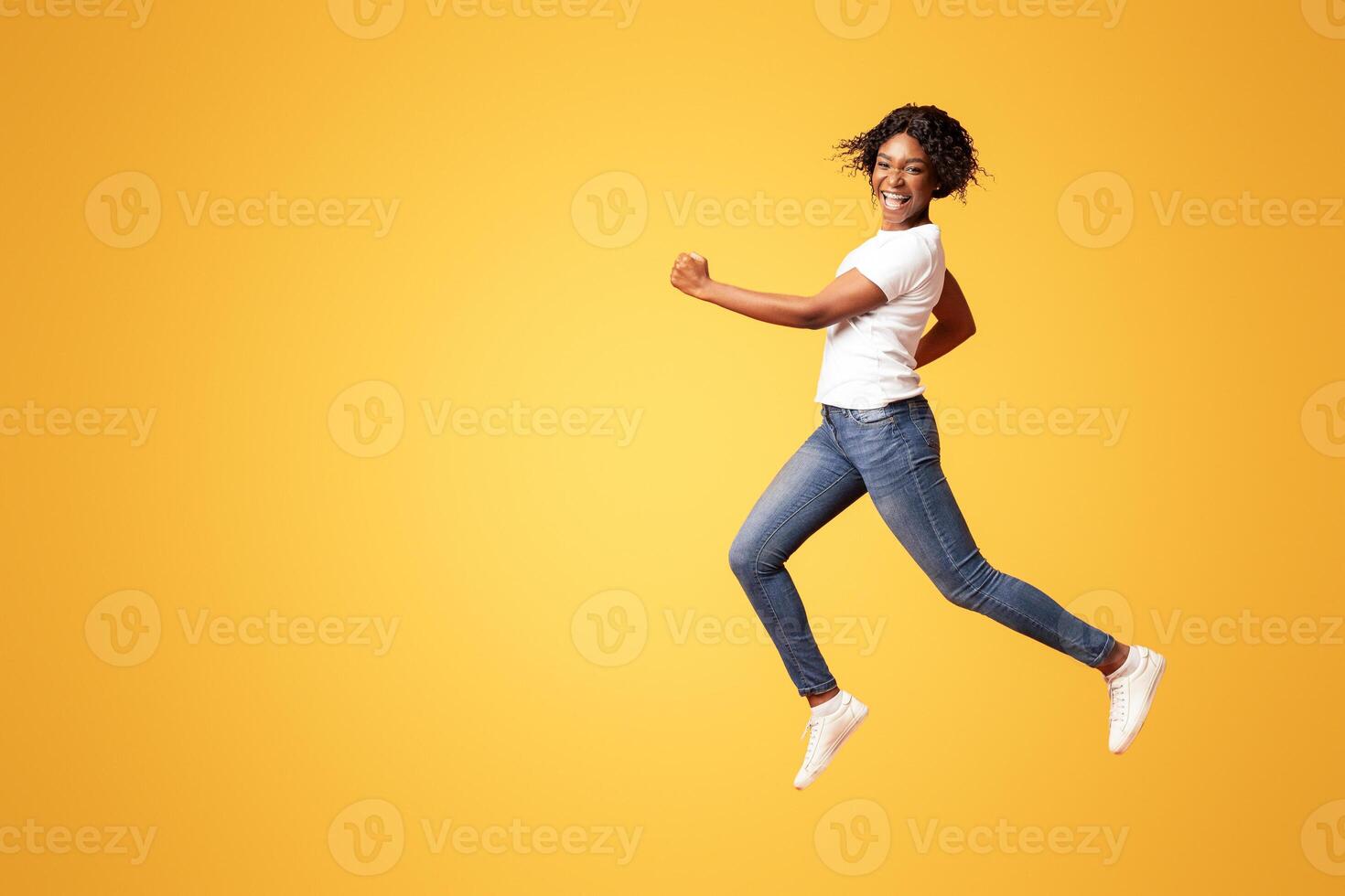 Active black lady in casual outfit running over orange studio background, full length shot. Cheerful african american young woman in jeans and white t-shirt having fun on her own photo