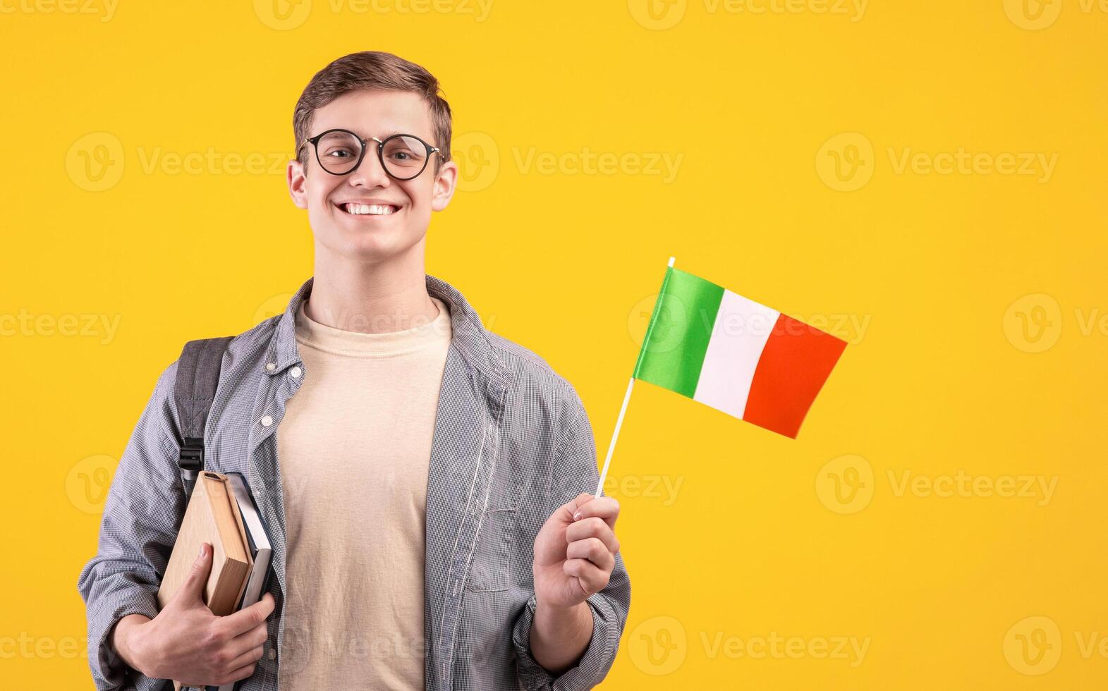 Love of country, education and travel, exchange student and new pupil. Young smiling guy in glasses with backpack holds books and small flag of Italy, isolated on beige background, studio shot photo