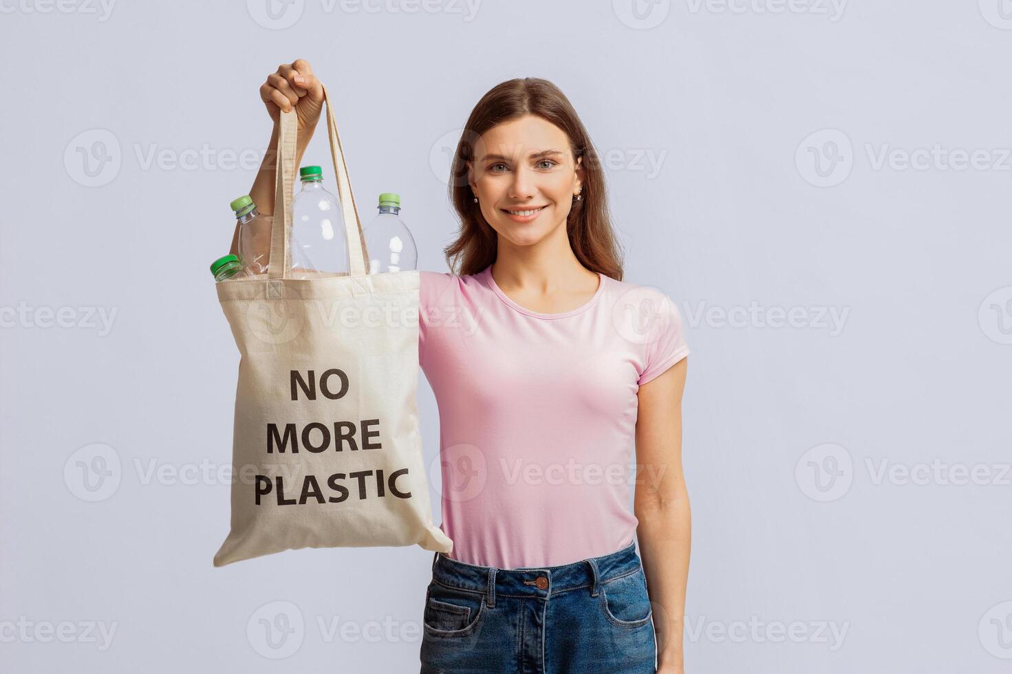 A young woman stands against a light background, cheerfully holding a reusable bag filled with plastic bottles. She advocates for sustainable recycling practices and an eco-friendly lifestyle. photo