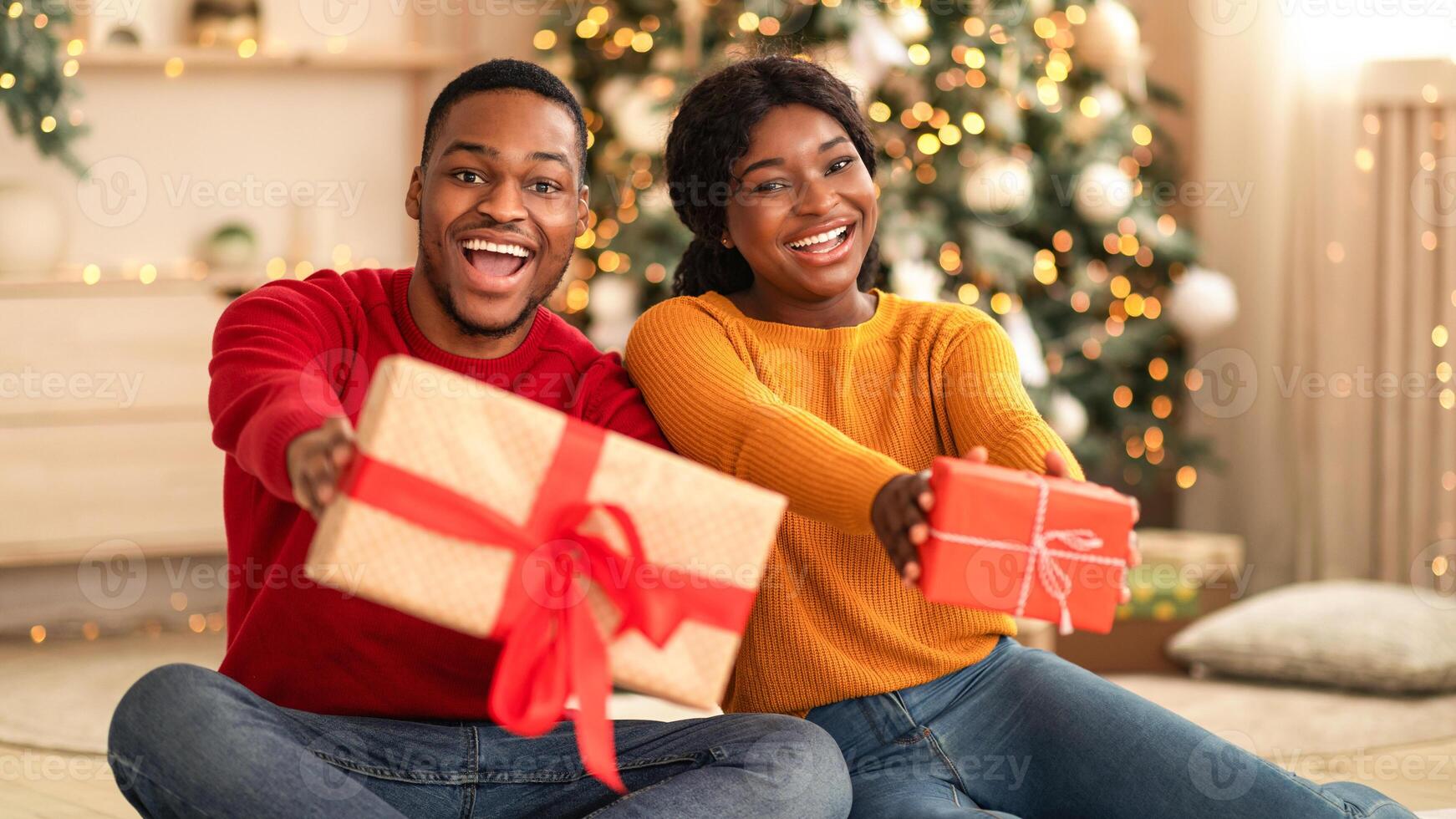 Fulfillment of wishes in New Year at home during lockdown. Laughing millennial african american man and woman show boxes with gifts and ribbons in living room interior with Xmas tree and garlands photo