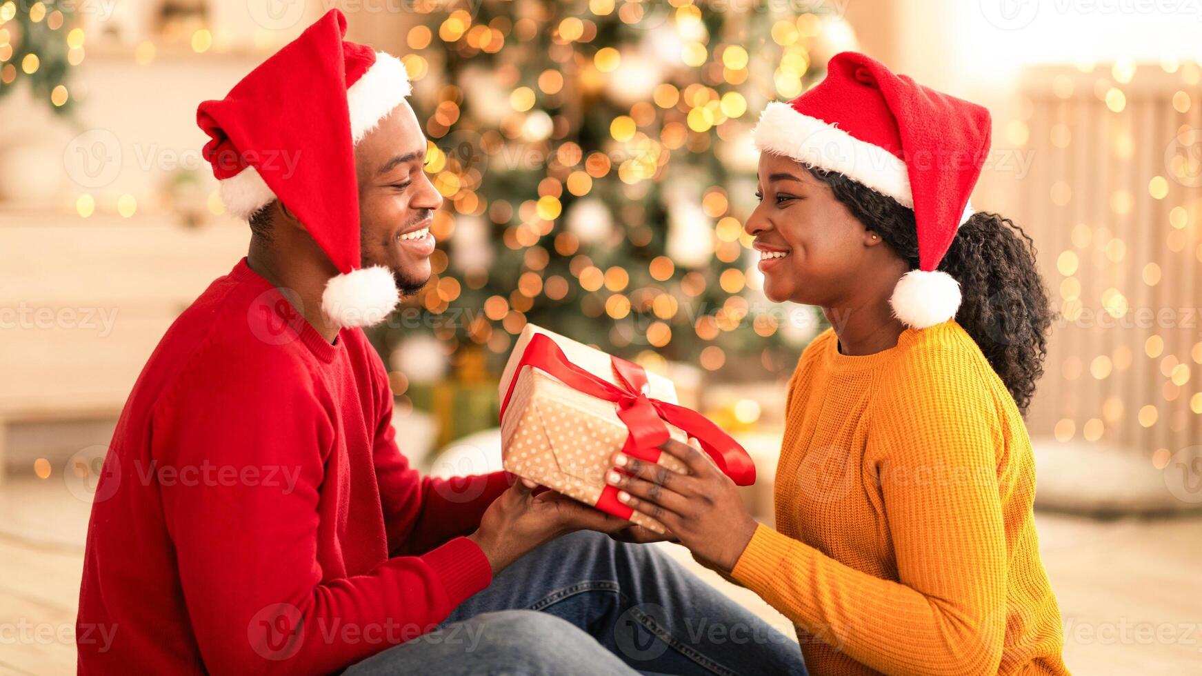 Romantic, New Year and Xmas together. Happy young african american man and woman in Santa hats exchange gifts in interior with Christmas tree and living room decor with glowing garlands, profile photo