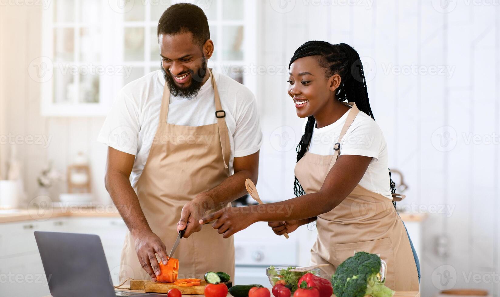 Happy African American Spouses Using Laptop Computer While Cooking In Kitchen Together, Searching Recipe In Internet, Smiling Black Couple Preparing Healthy Food For Dinner At Home, Copy Space photo
