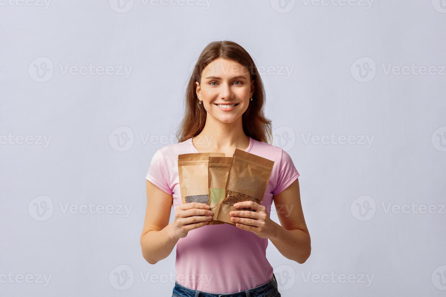 Eco Storage Concept. Smiling millennial lady holding set of brown kraft paper doypack bags with weight groceries, using biodegradable ziplock packs for food storing, standing over light background photo