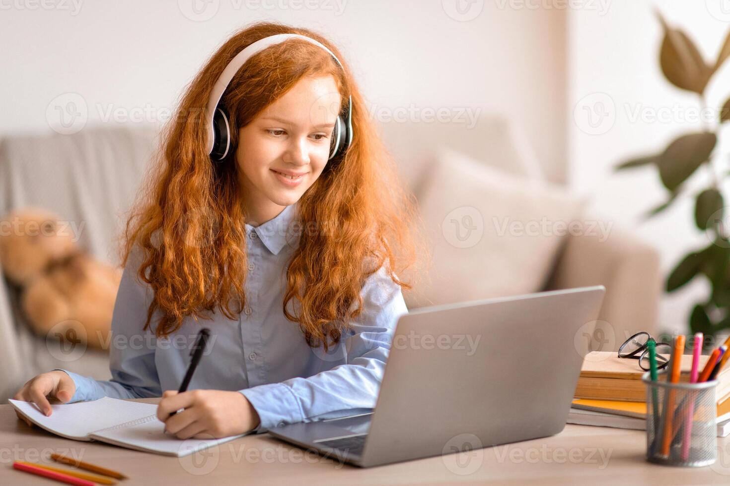 Doing Homework. Smiling young teen girl sitting at desk with pen, writing and taking notes in notebook, using and looking at laptop computer. Cheerful pupil in headphones watching online course photo