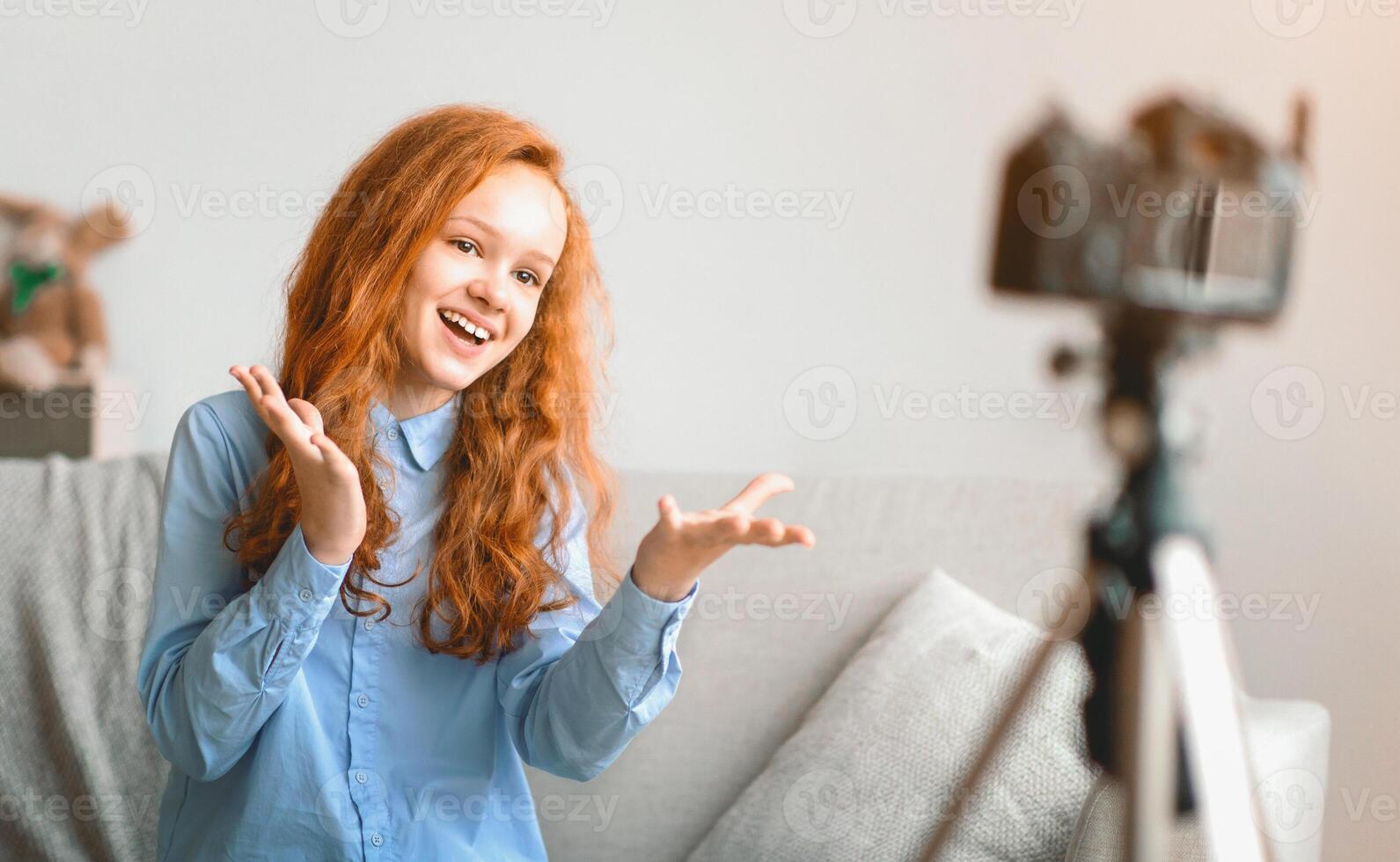 Blogging Concept. Smiling red-haired teen girl recording lifestyle blog talking to camera on tripod, sitting on the couch at home. Happy young influencer filming review for her internet channel photo