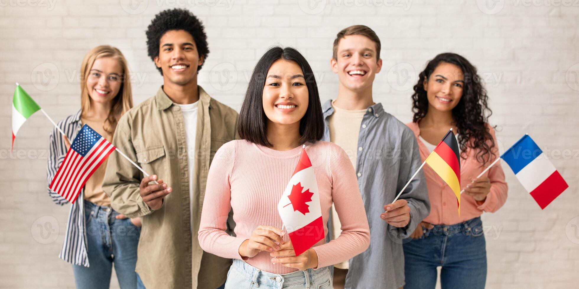 Young students of language school celebrate holiday. Happy millennial peoples of different nationalities have fun on study and hold little flags on brick white wall background, studio shot, free space photo