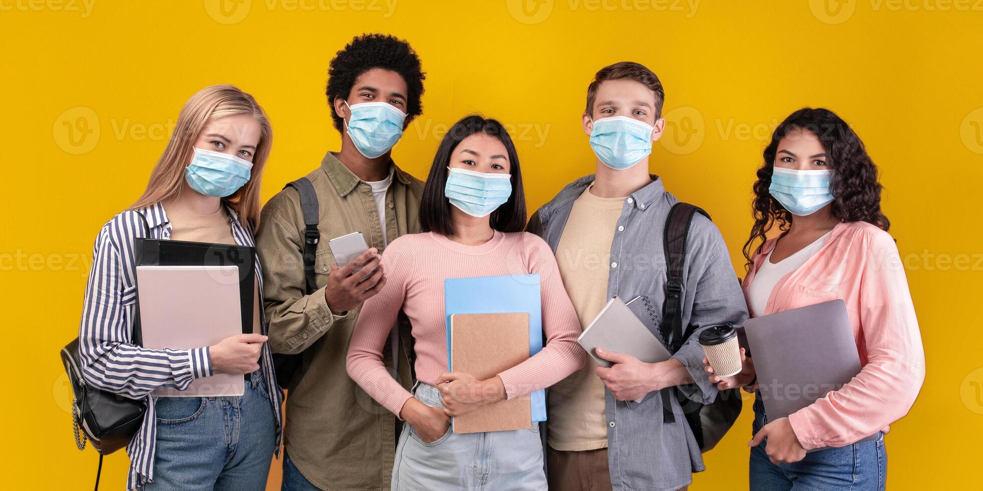 Study abroad and student exchange during covid-19 lockdown. Modern cheerful teens multiracial students with notepads, gadgets, cups of coffee looking at camera, isolated on orange background photo