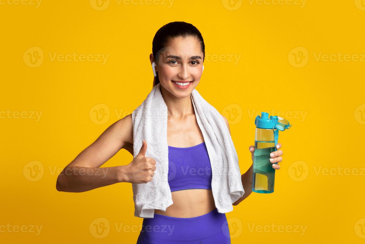 Stay Hydrated. Portrait of happy smiling female fitness instructor in wireless earphones holding bottle with fresh water and showing thumbs up sign gesture, posing isolated on orange studio background photo
