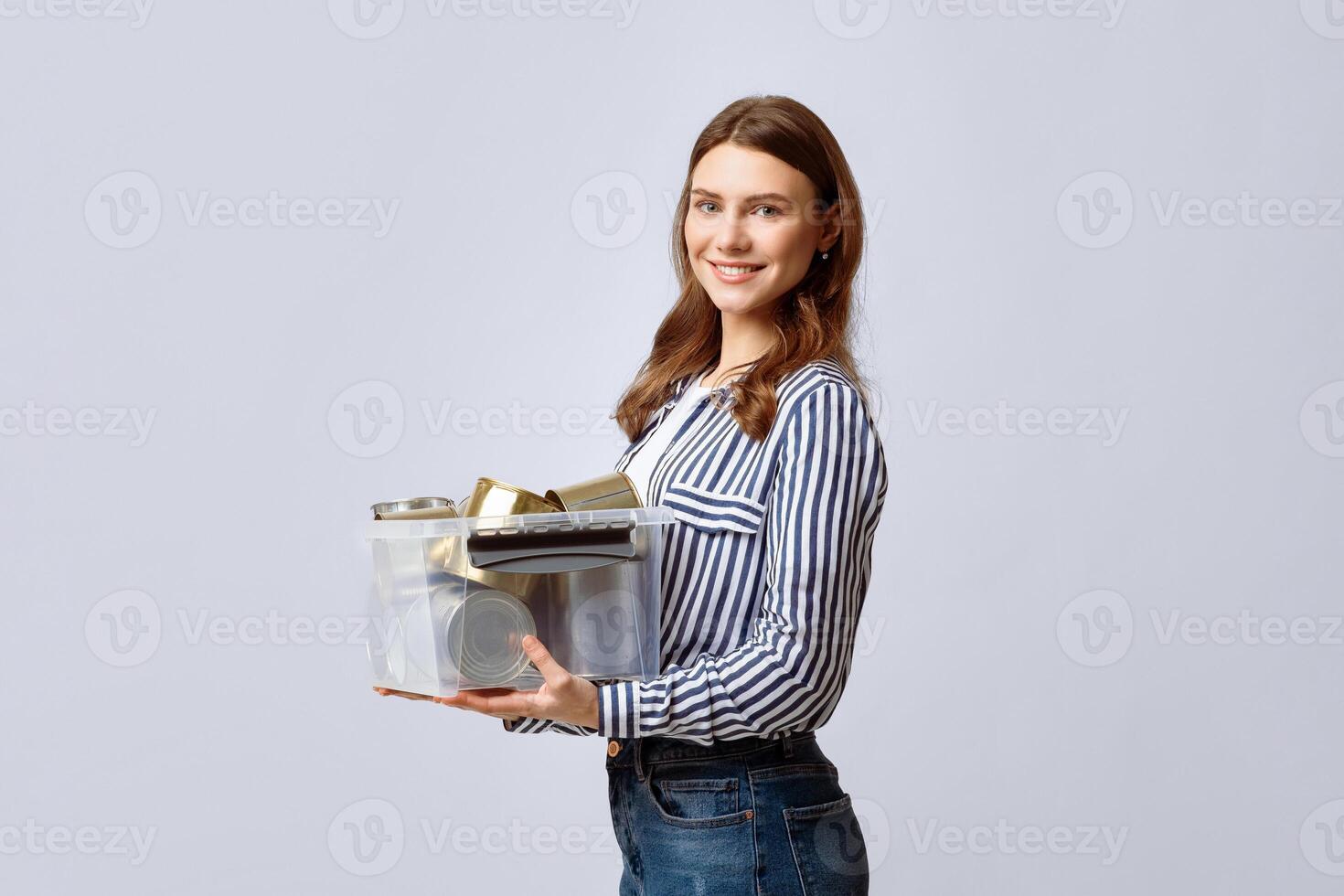 A young woman stands indoors, smiling broadly while holding a transparent container filled with recyclable materials. She showcases a commitment to eco-friendly practices and sustainability. photo