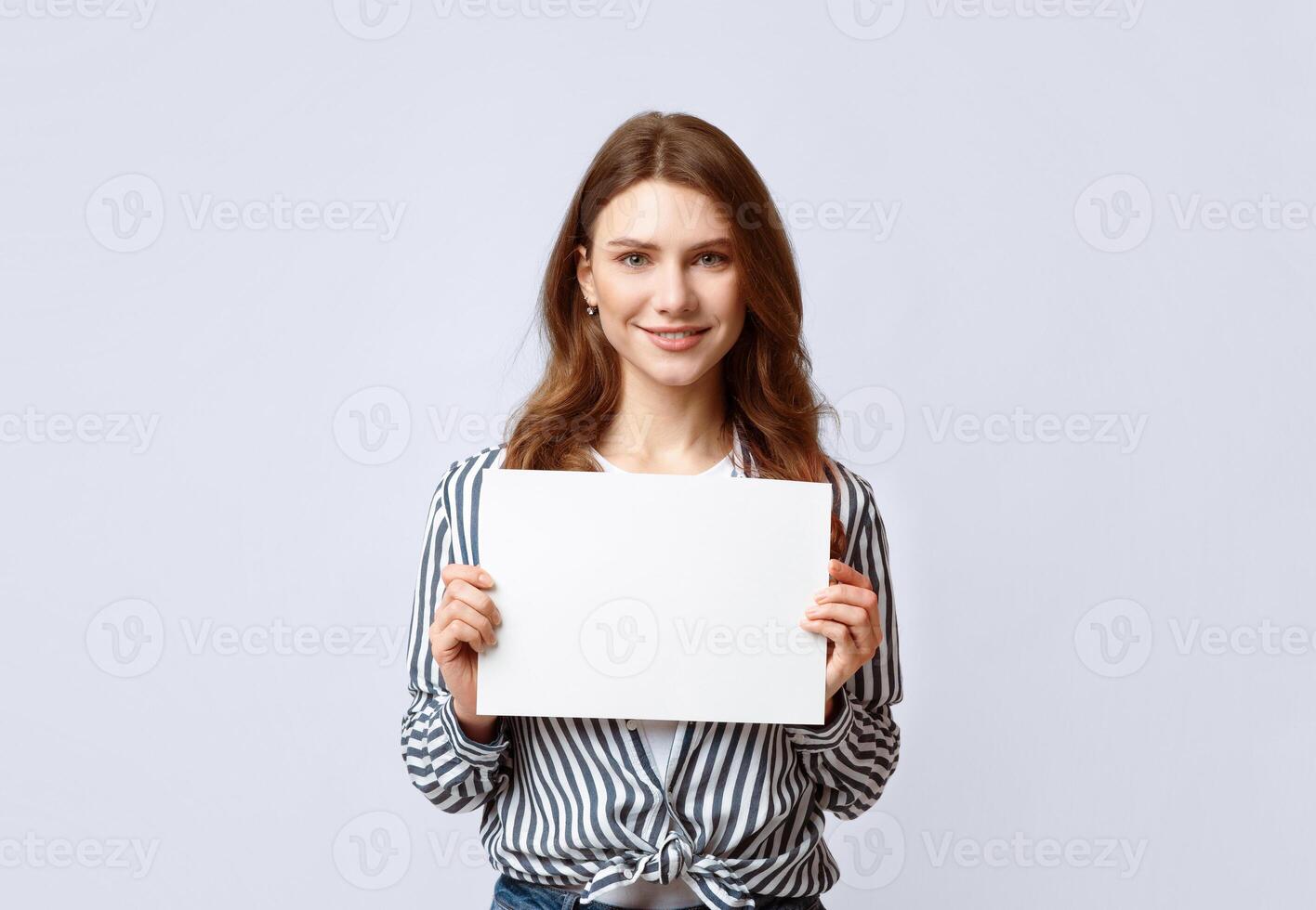 Portrait of beautiful woman holding white blank advertising board, smiling millennial lady demonstrating empty banner with copy space for your text or design, standing on gray background in studio photo