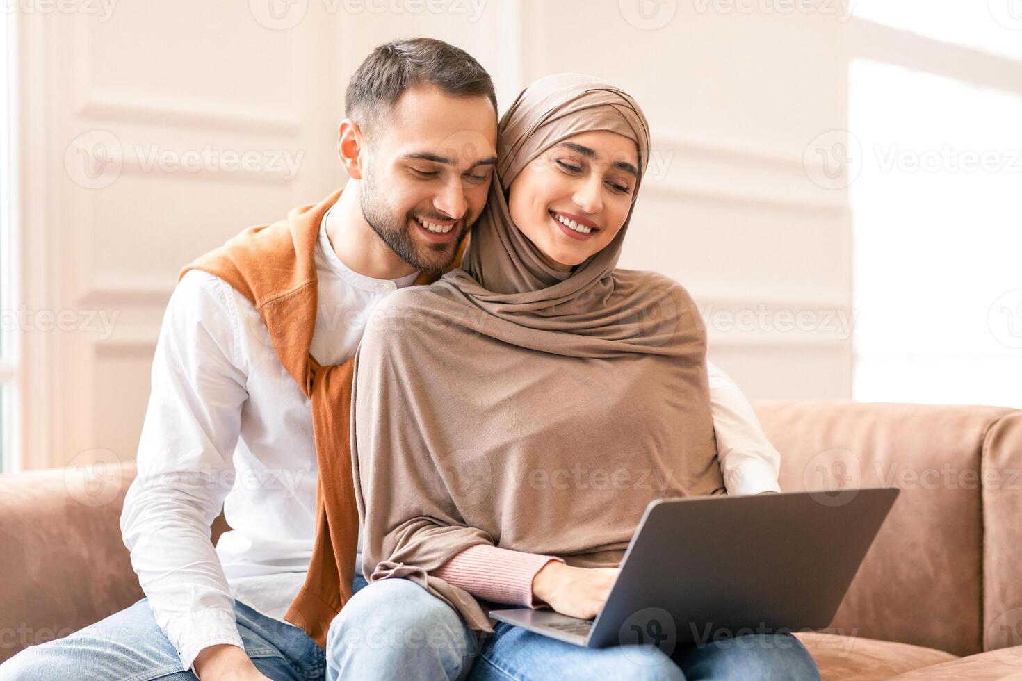 Modern Muslim Couple Watching Movie Online On Laptop Computer Sitting And Hugging On Couch, Relaxing At Home On Weekend. Lovely Arab Spouses Browsing Internet Together photo