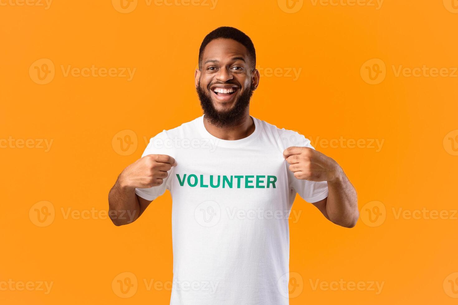Volunteer's Portrait. Happy Black Activist Man Showing His T-Shirt With Volunteer Print Inscription On Chest Standing On Yellow Studio Background. Volunteering Job Concept photo