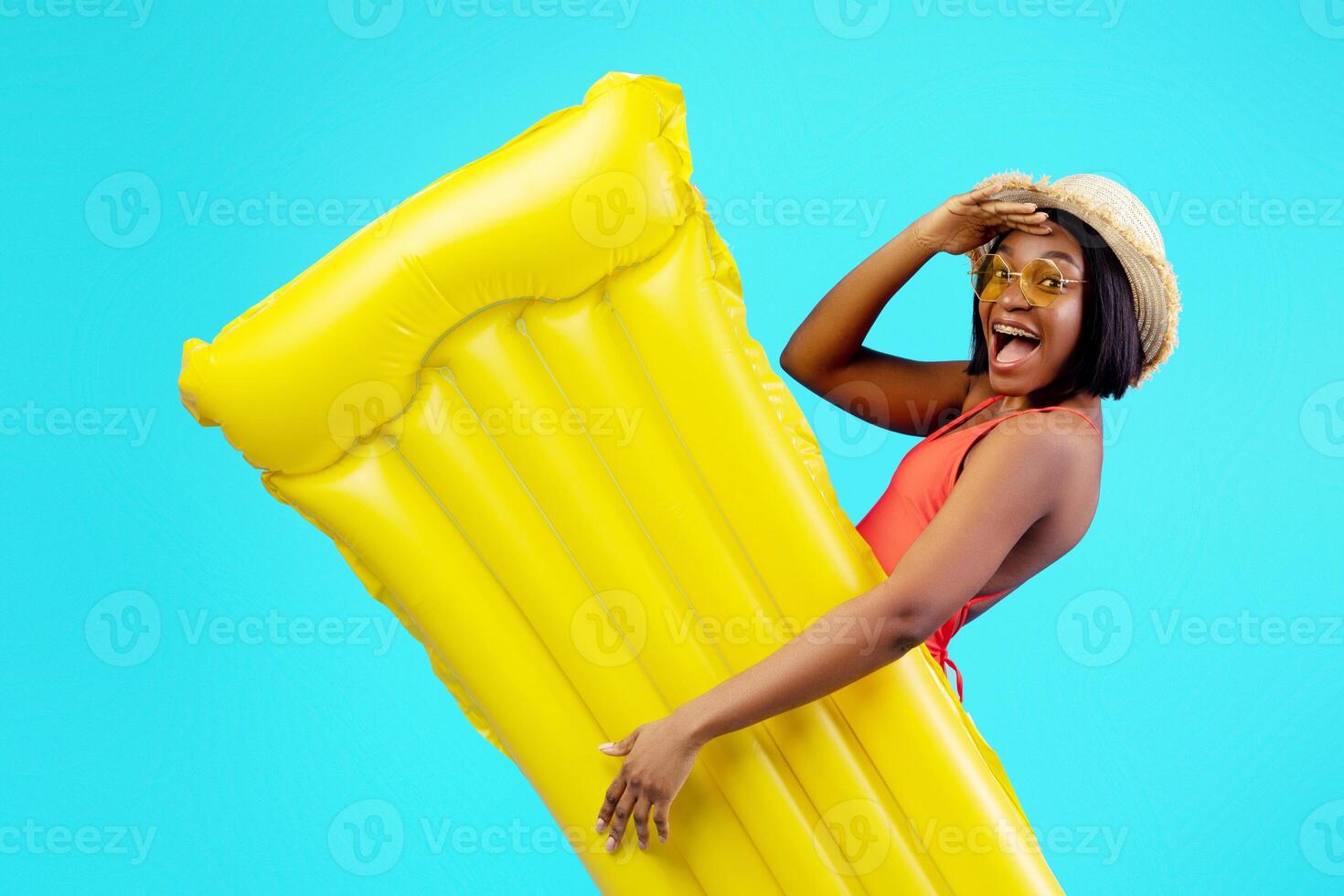 Millennial black woman in swimsuit holding yellow inflatable lilo on blue studio background. Young African American lady with pool float having tropical summer vacation or beach party photo