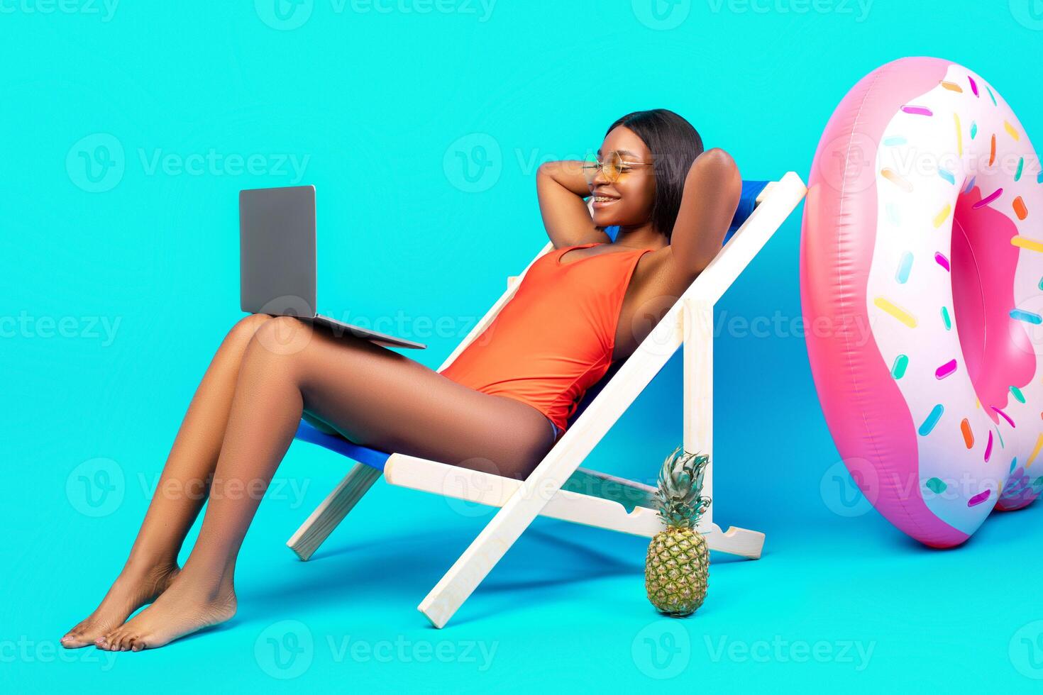 Relaxed black lady chilling in lounge chair with laptop computer, communicating online from tropical paradise, watching good movie on summer vacation, blue studio background. Copy space photo