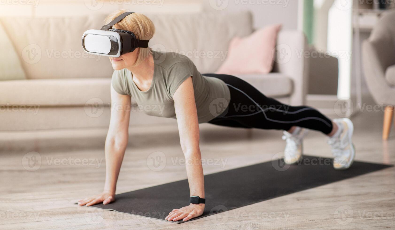 Exercising with virtual reality concept. Mature woman in VR headset standing in plank pose at home. Strong senior lady using modern technologies for domestic yoga workout photo