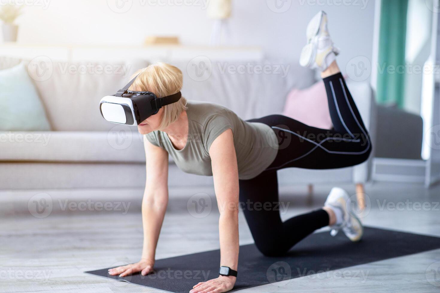 Sports and virtual reality concept. Senior woman in VR headset exercising at home. Athletic mature lady using modern technologies for domestic workout, doing fitness indoors photo