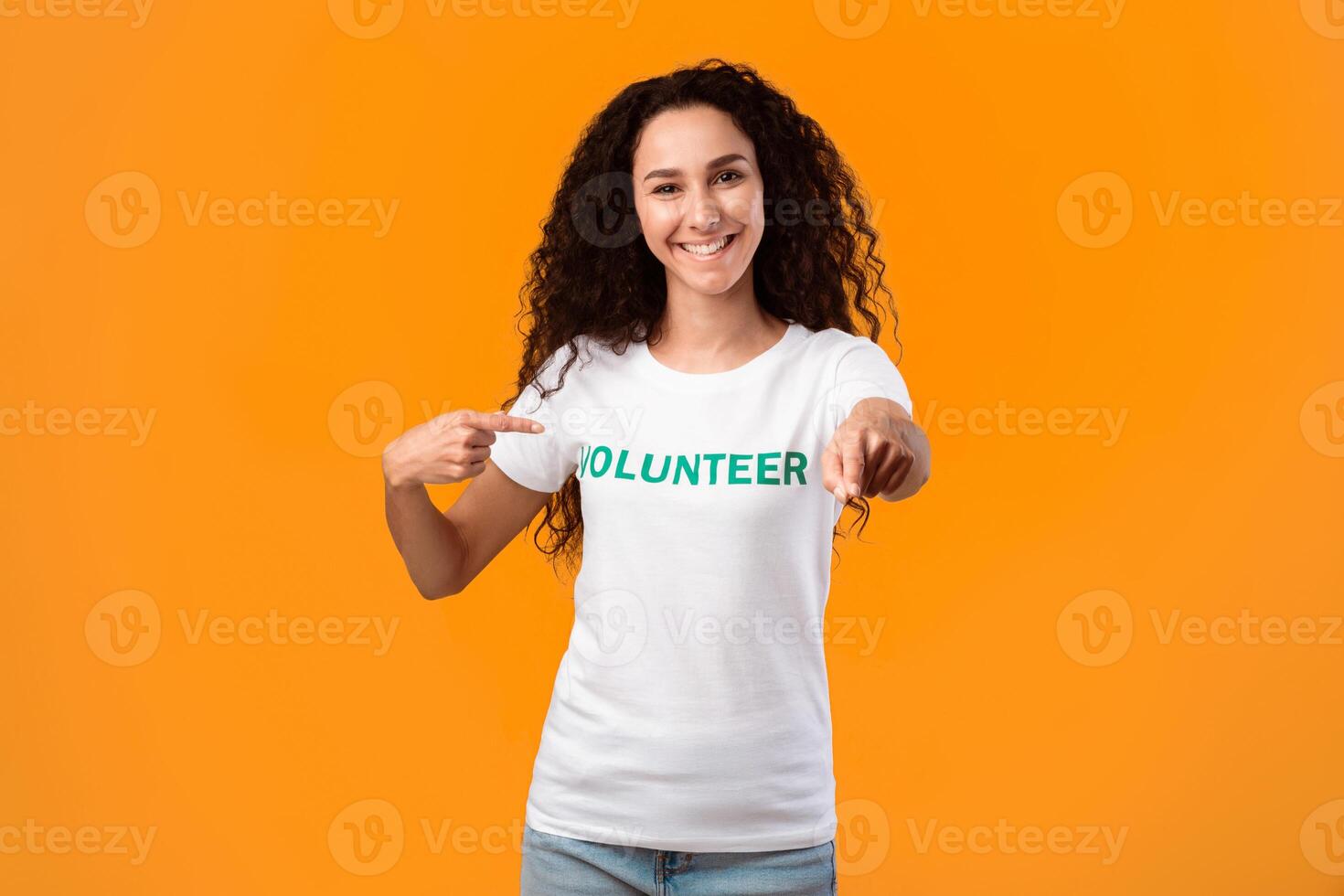 I'm a volunteer concept. Joyful female activist pointing at t-shirt uniform and at camera posing in studio over yellow background. Volunteering career, charity work photo
