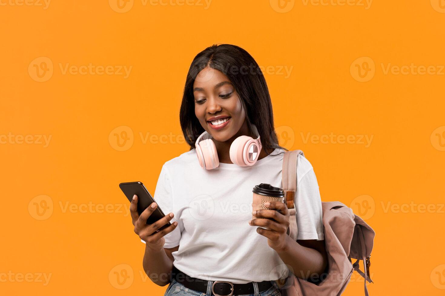 Stylish young black lady student with backpack and wireless headset holding mobile phone and coffee to go, having break while studying, chatting with friends, orange studio background, copy space photo
