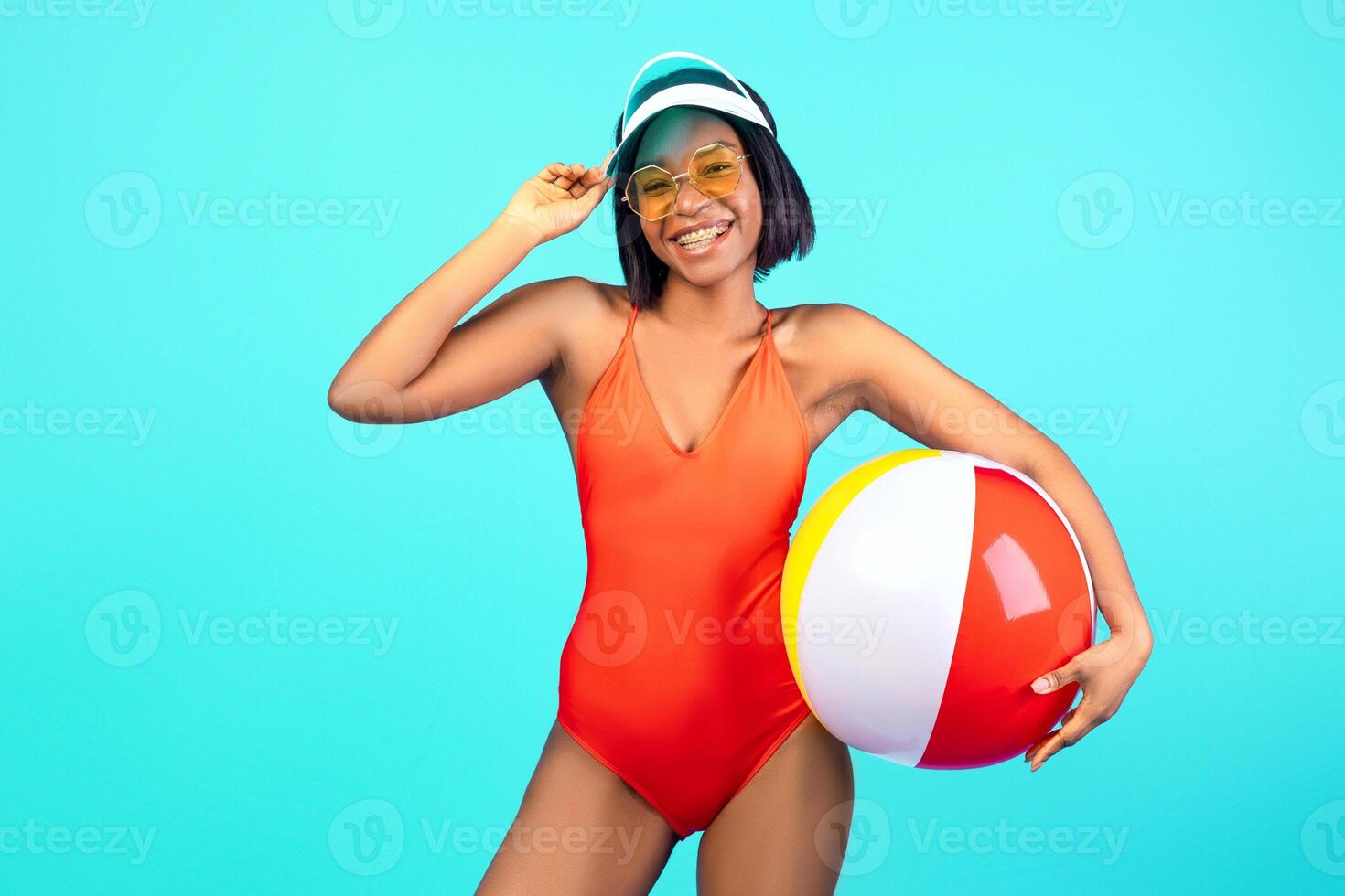 Carefree black woman in swimsuit posing with inflatable beach ball, smiling and looking at camera on blue studio background. Portrait of happy young lady chilling on summer beach vacation photo