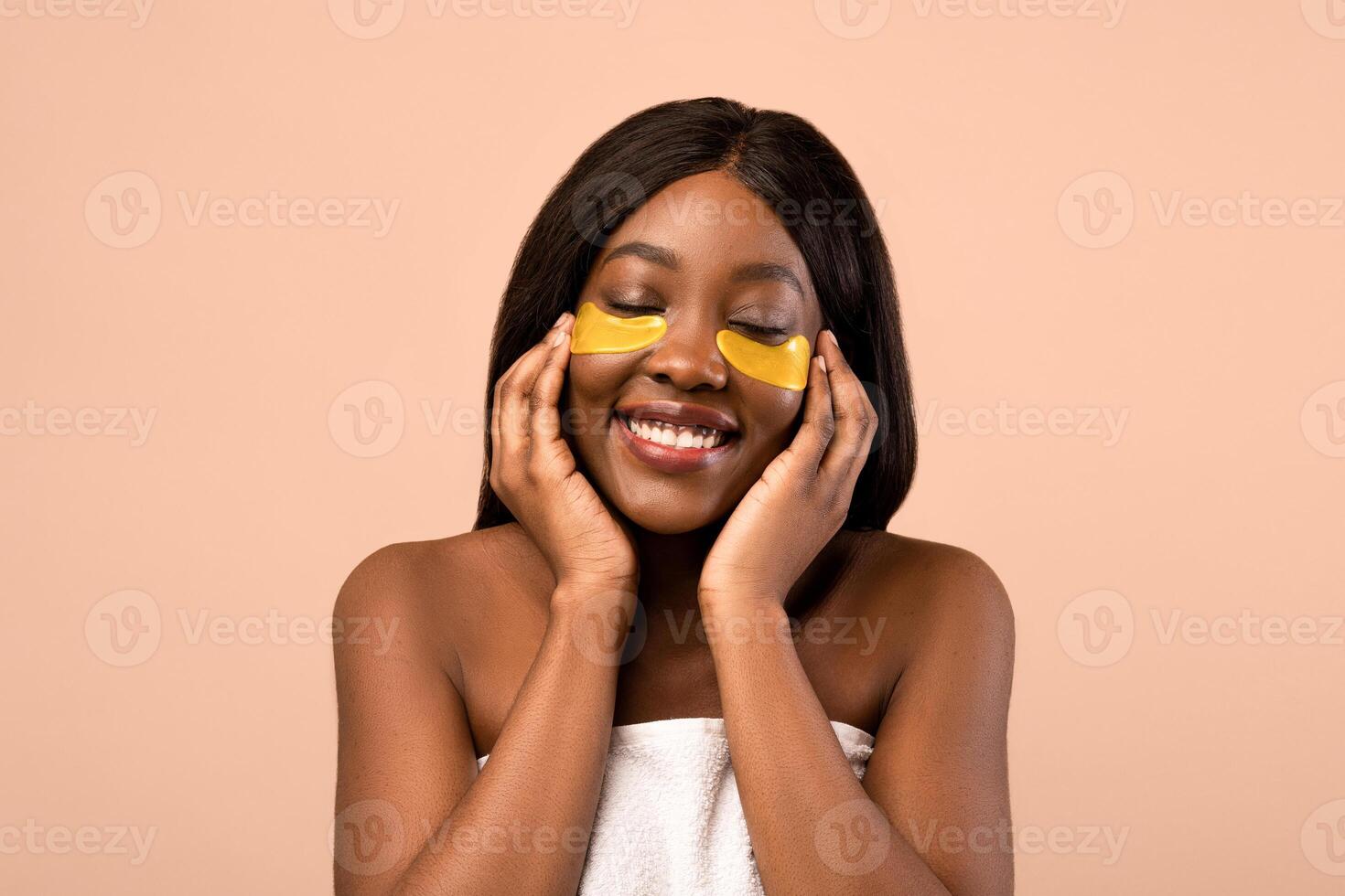 Closeup of pretty joyful young black lady with closed eyes applying golden hydrogel eye patches over beige studio background, copy space. Anti-aging, against lines, wrinkles eye care concept photo