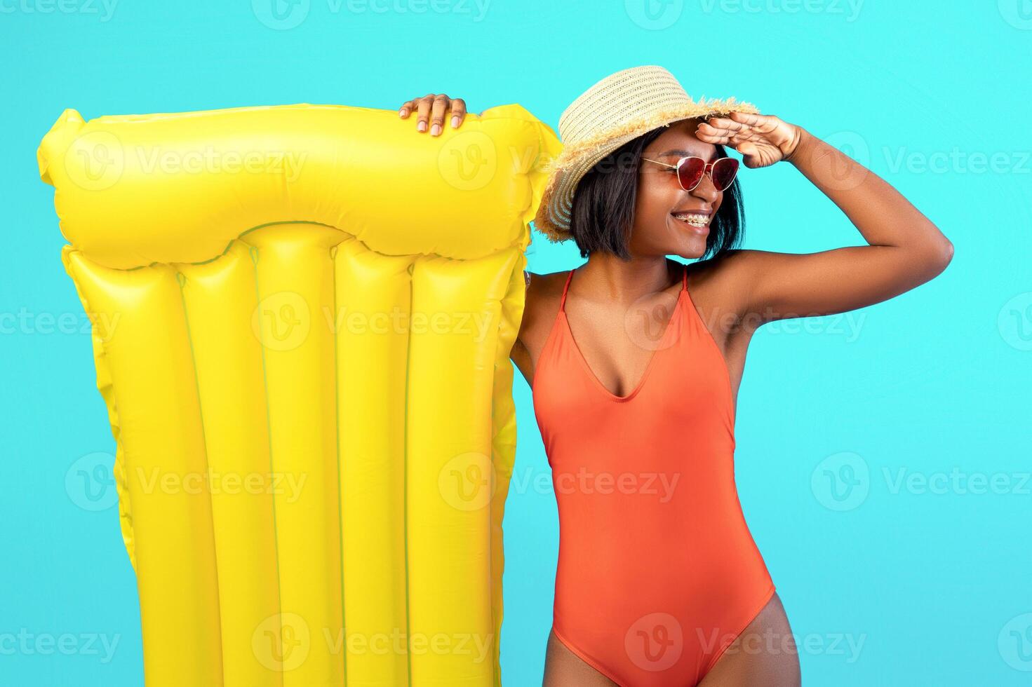 Millennial black lady in swimsuit with inflatable lilo holding hand above her eyes, looking into distance on blue studio background. African American woman with pool float having great summer vacation photo