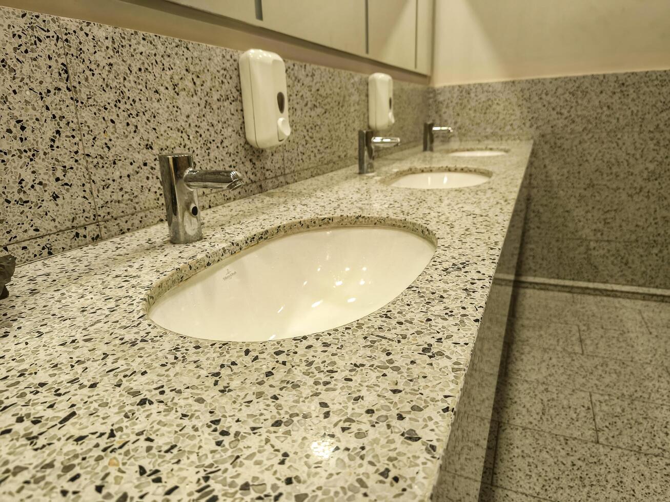 A bathroom with three sinks and a soap dispenser. The sinks are white and the counter is made of granite photo