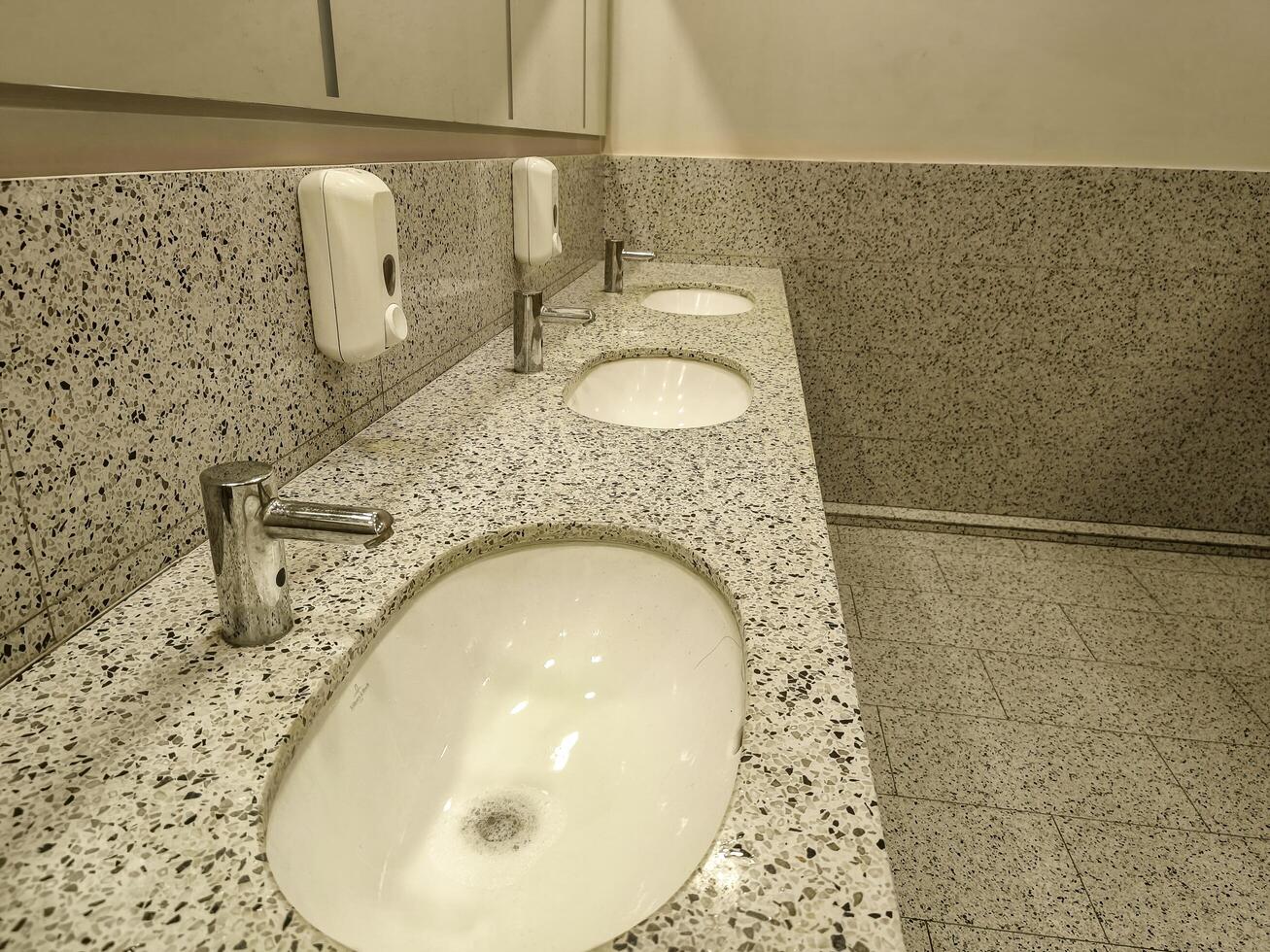 A bathroom with three sinks and a soap dispenser. The sinks are white and the walls are white photo