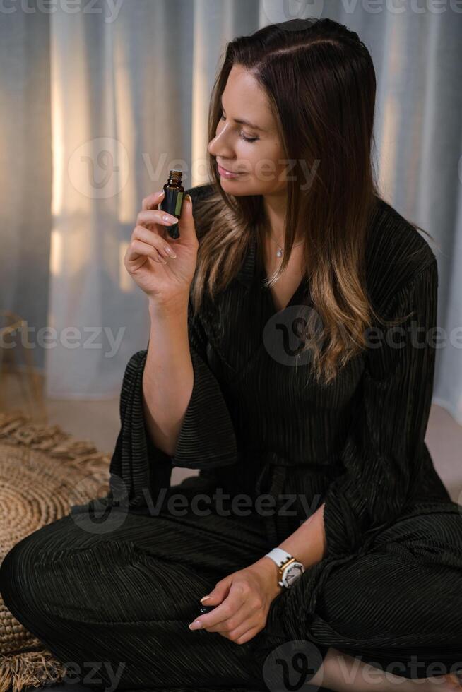Woman holding essential oil bottle while sitting calmly on the floor in a serene indoor environment during a relaxing moment of self-care and mindfulness practice at dusk photo