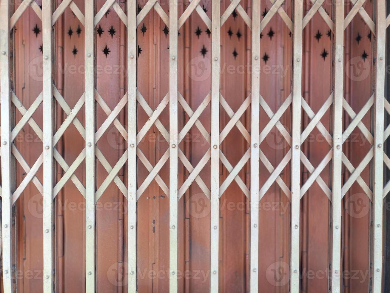 Vintage metallic folding gate background texture detail for protection. Old residential security shutter design against brown wooden wall surface. Collapsible steel door protector with classic photo