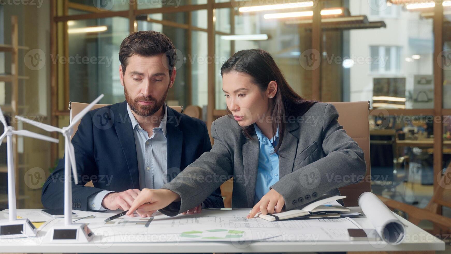 Japanese businesswoman inspector Caucasian businessman speak wind turbine model eco implementation renewable project document blueprint electricity engineering indoors office multinational meeting photo