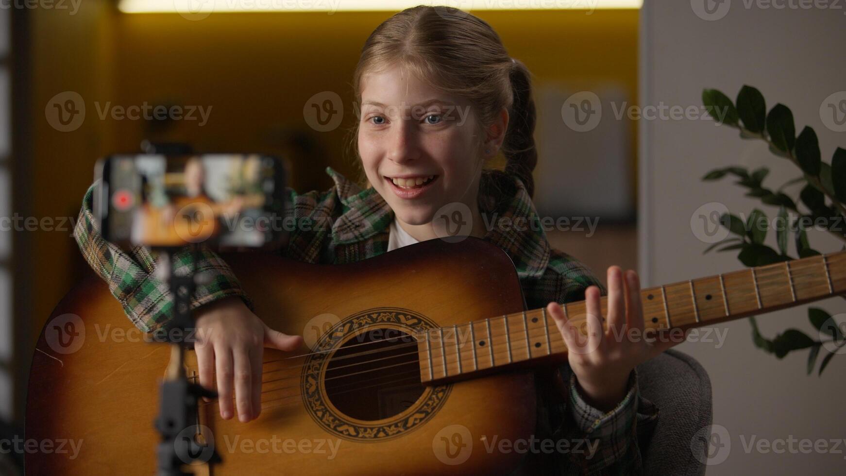 Creative little girl blogger Caucasian teaching how to play guitar record mobile phone camera tripod learning playing music influencer child kid filming tutorial song musical instrument at home photo