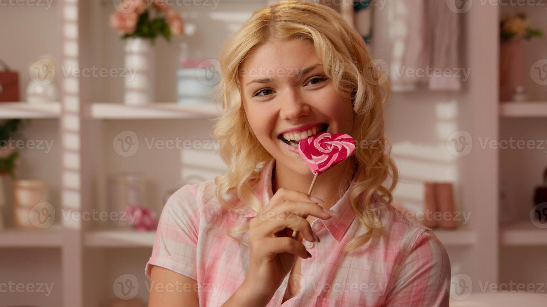 Caucasian girl female teenager woman in pink candy glamour bathroom holding striped candy red lollipop in heart shape biting eating yummy sugar snack lollypop looking at camera indoors bite eat happy photo