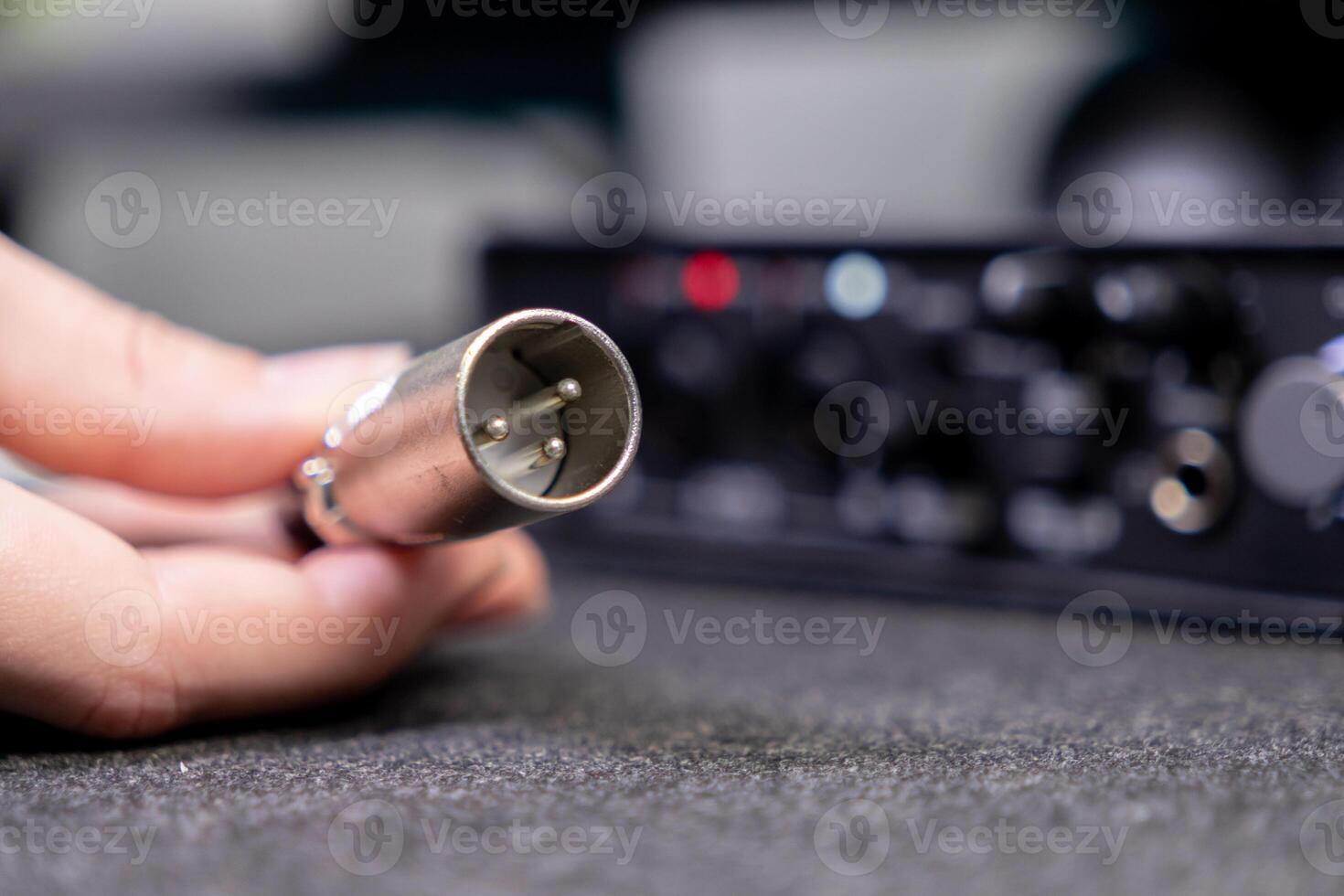 Human hand holds an XLR audio cable connector, poised for connection to a blurred professional sound interface, showcasing audio engineering and recording photo