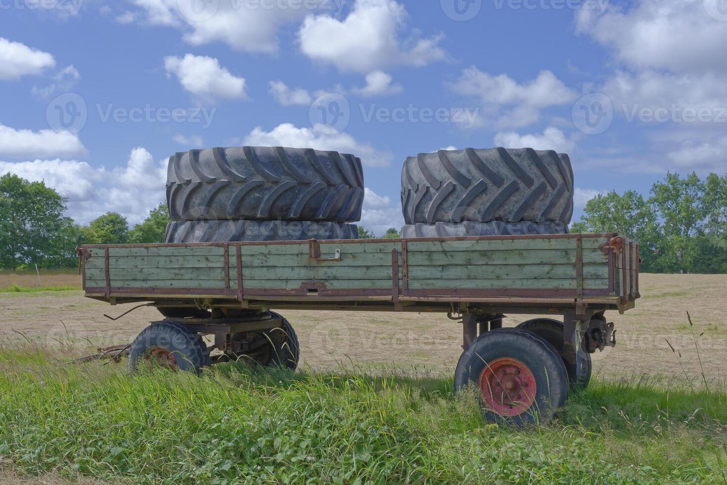At the edge of the field stands a large trailer with four large tractor tires under a blue sky photo