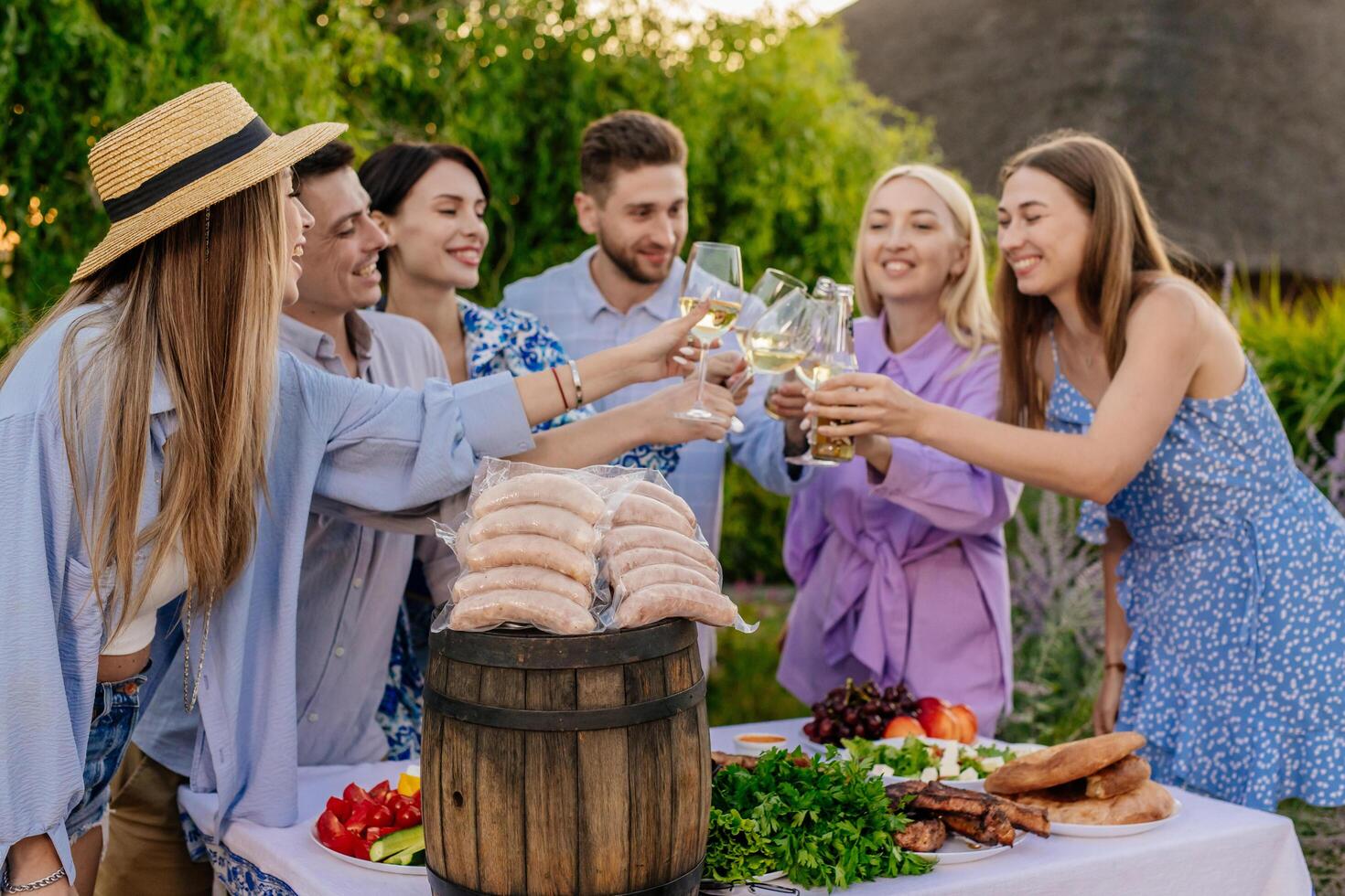Happy friends eating, drinking beer, and raising a toast at a BBQ dinner in nature, next to sausages for the barbecue. Concept food and friendship, promoting BBQ and outdoor lifestyle photo