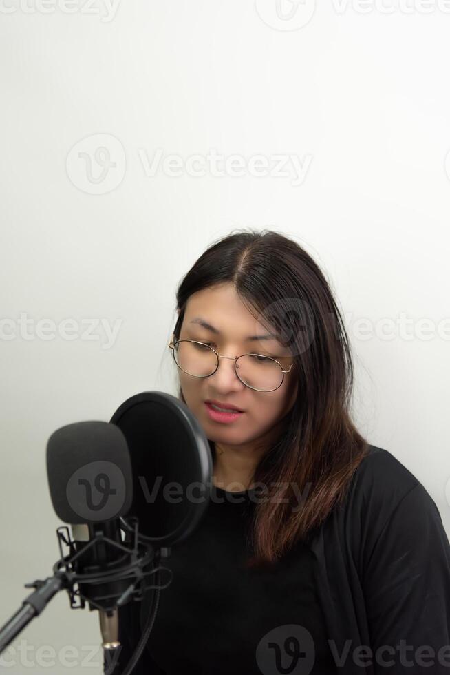 A woman with glasses and a microphone in front of a white wall photo