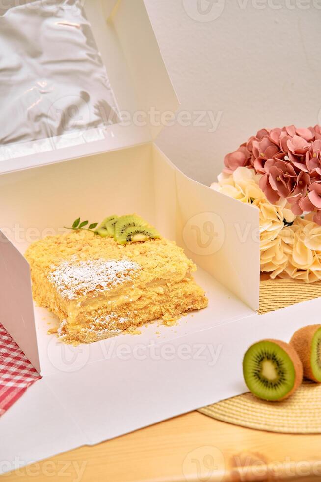 Luscious layered cake in white box with kiwi garnish and fresh flowers on wooden table photo