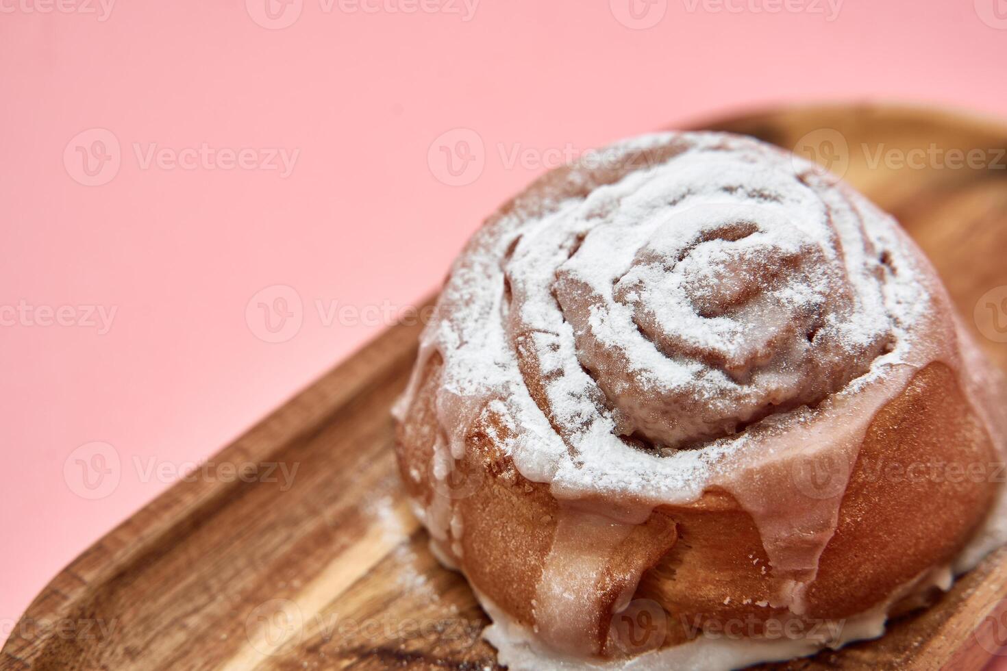 Cinnamon roll with frosting on wooden plate against pink background photo