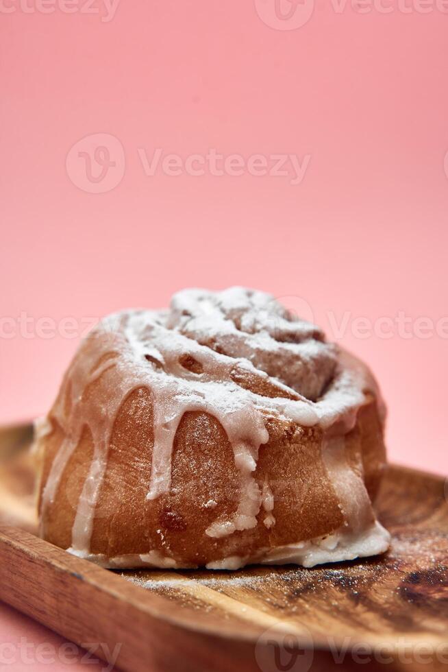 Cinnamon roll with icing on wooden platter against pink background photo