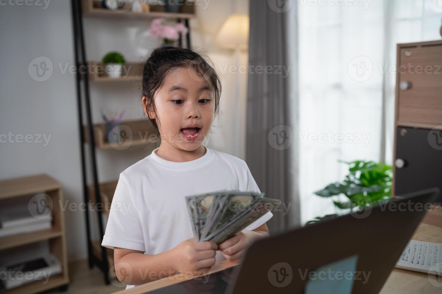 Happy child holding cash while using laptop at home, excited expression, indoor setting, finance education, youthful entrepreneur experience photo