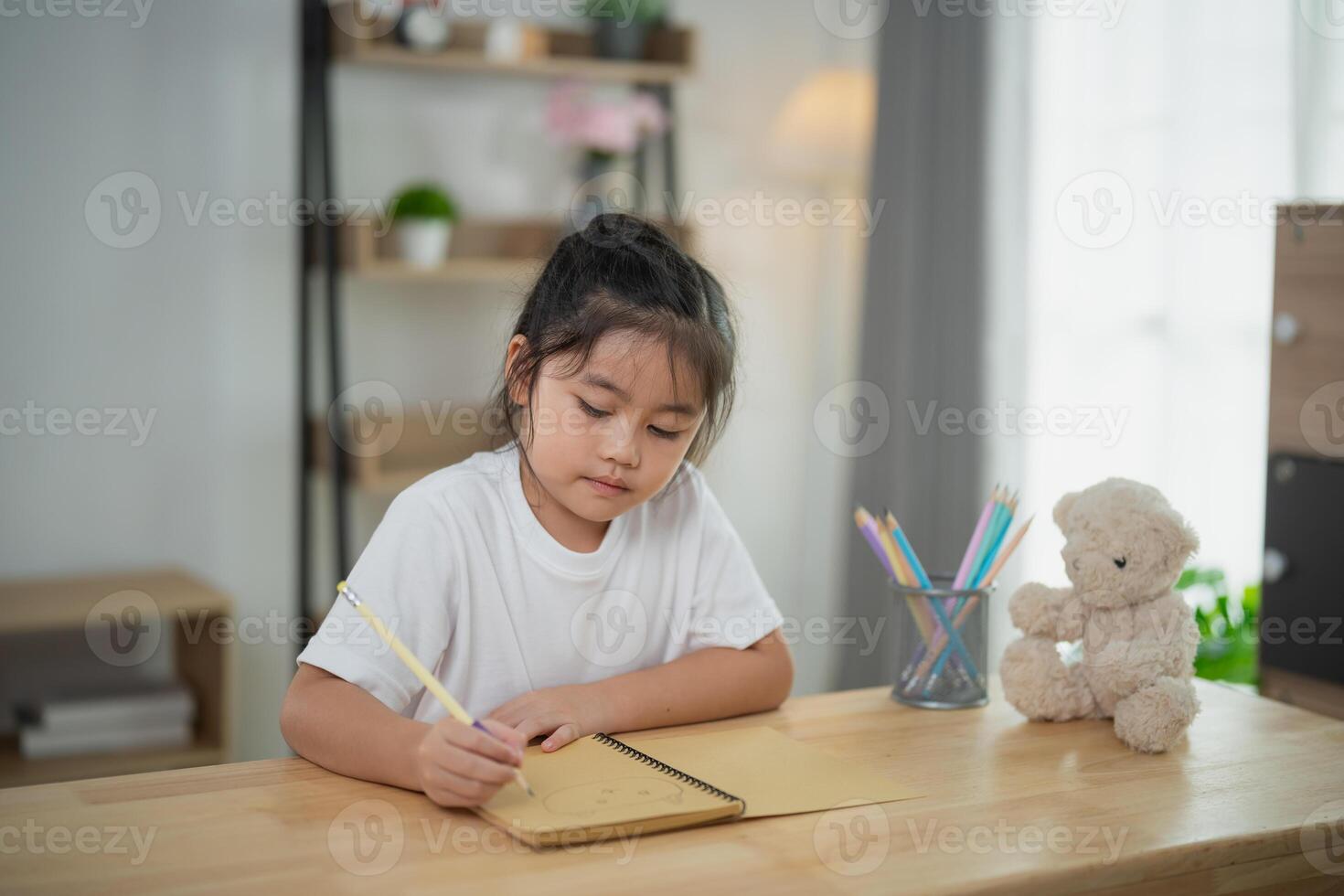Young girl engaged in creative writing or drawing in a cozy indoor setting with soft light, surrounded by a teddy bear and colorful art supplies on a wooden table photo