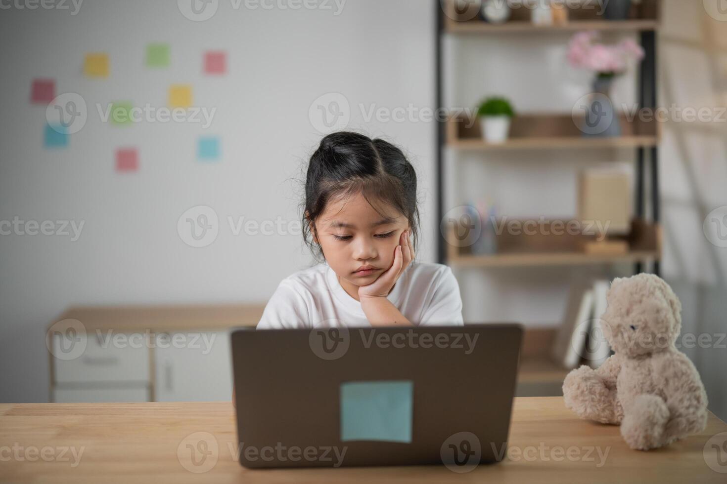 Young girl with thoughtful expression using laptop on wooden table in a cozy room with soft toys and colorful sticky notes on the wall. photo