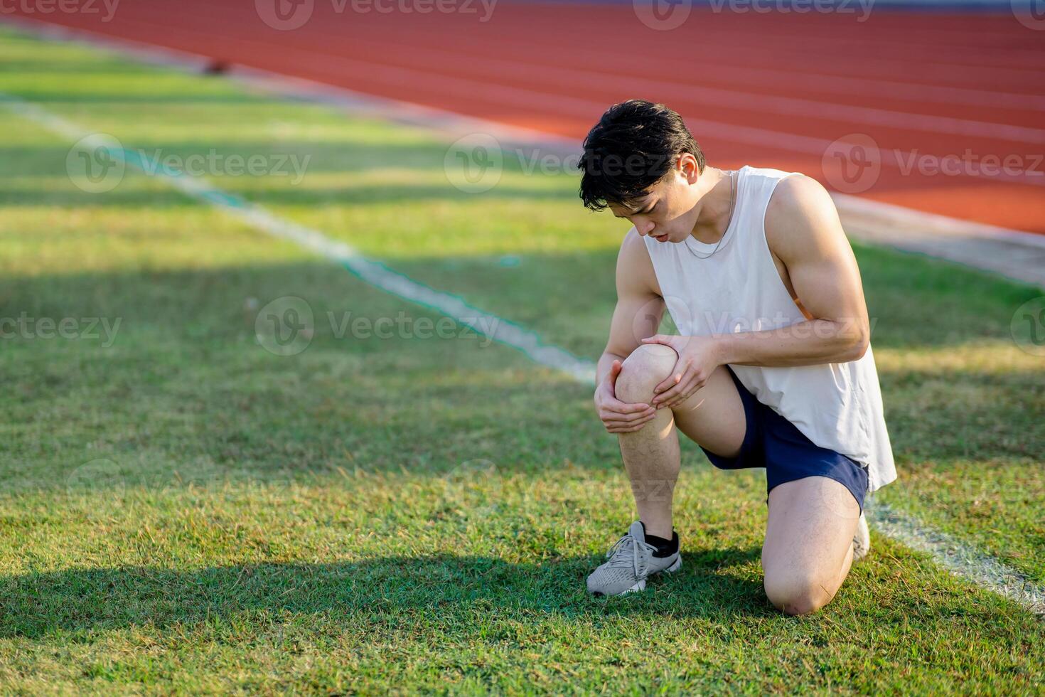 White tank top asian man is looking at his knee while crouching on grass field. Pain or injury. photo