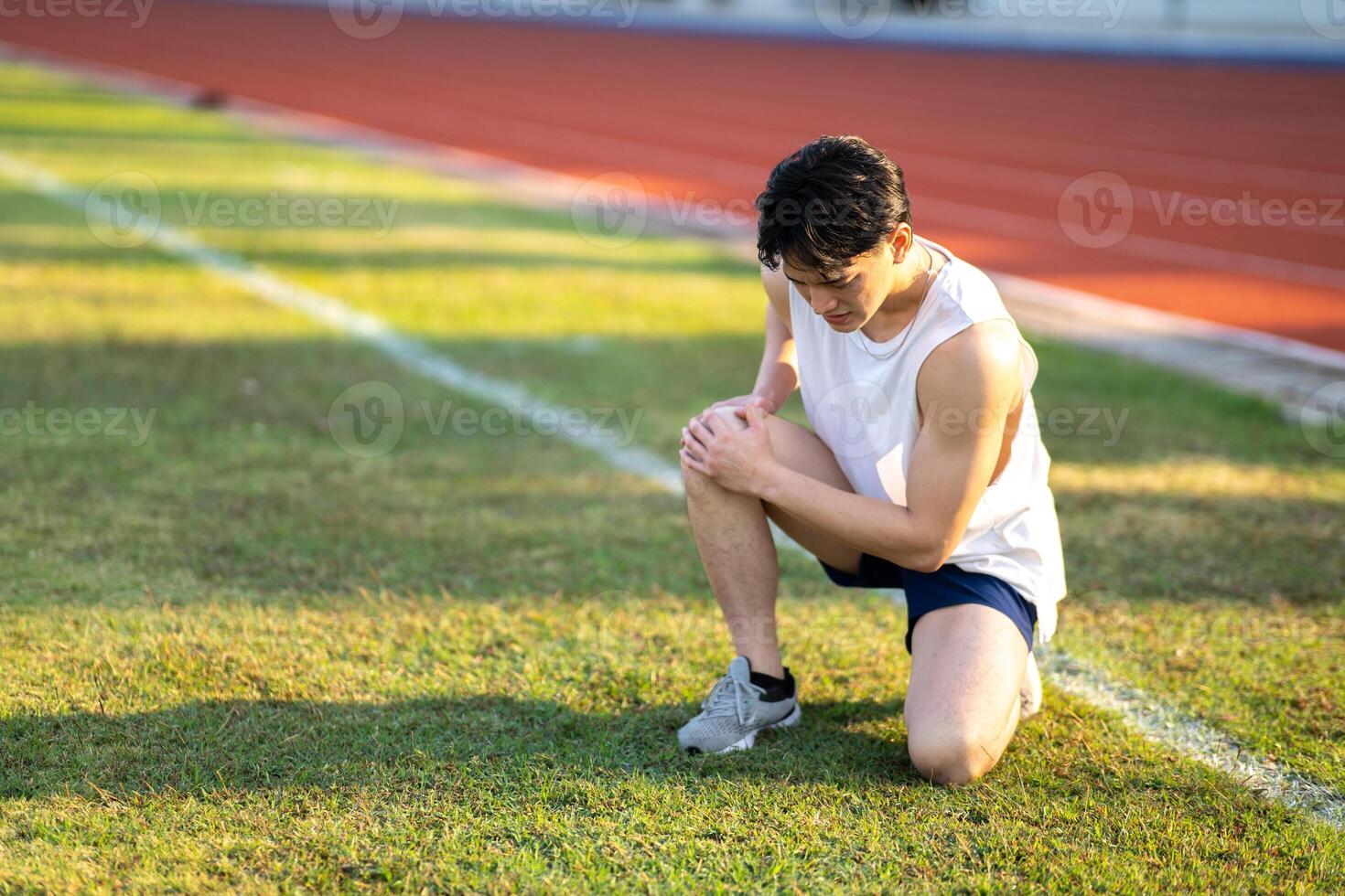 White tank top asian man with hands on his knee is crouching on grass field. Pain or injury. photo