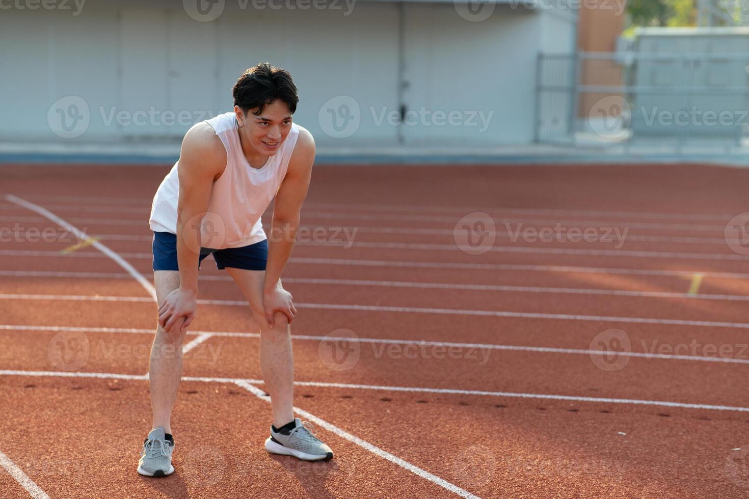 Asian man in white tank top looking ahead while standing with hand on knee, take a rest after run. photo