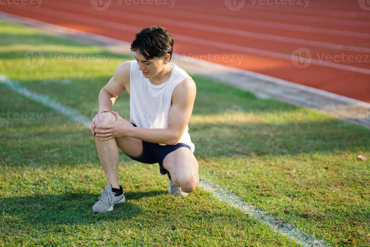 White tank top asian man is holding his knee while crouching on grass field. Pain or injury. photo