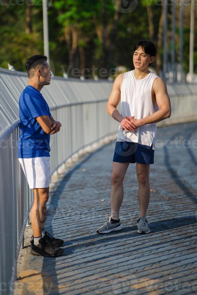 Two asian men or male friends talking together while standing at sports stadium fence or railing. photo